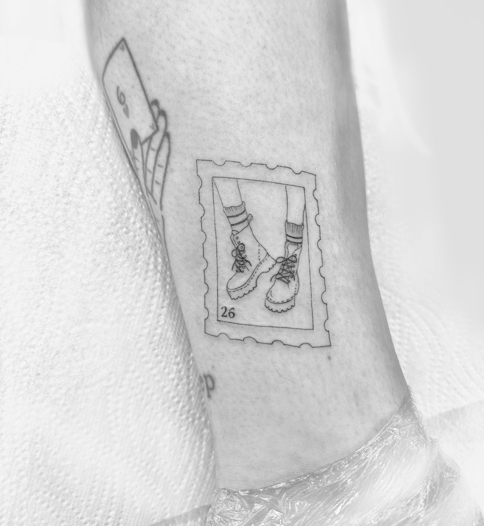 Fine line tattoo of a postage stamp showing two laced boots, with the number 26