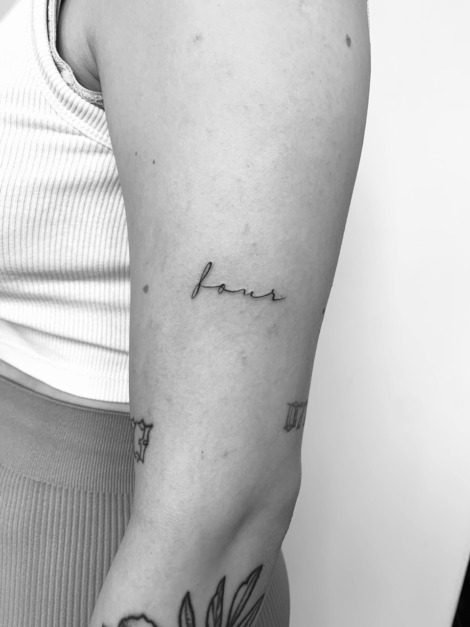 Fine line tattoo of the word four in handwritten script on an upper arm